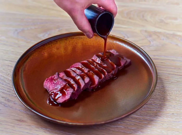 A server's hand drizzles steak sauce over beef slices arranged on a decorative plate, set against a neutral wooden background