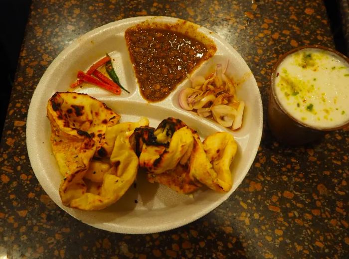 A divided plastic plate featuring crumbled, buttery naan, spiced pickled onions, hearty chickpea stew with peppers, and a refreshing cup of lassi.