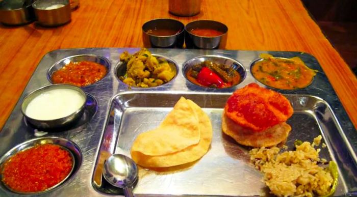 A colorful spread of a traditional thali features an array of small dishes, accompanied by flatbread, a pastry, and a serving of rice, all arranged on a tray.