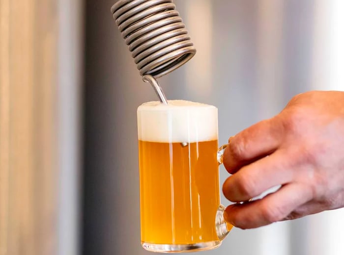 A hand grasps a full beer mug with a thick foam cap, positioned under a metal tap featuring a prominent spiral handle