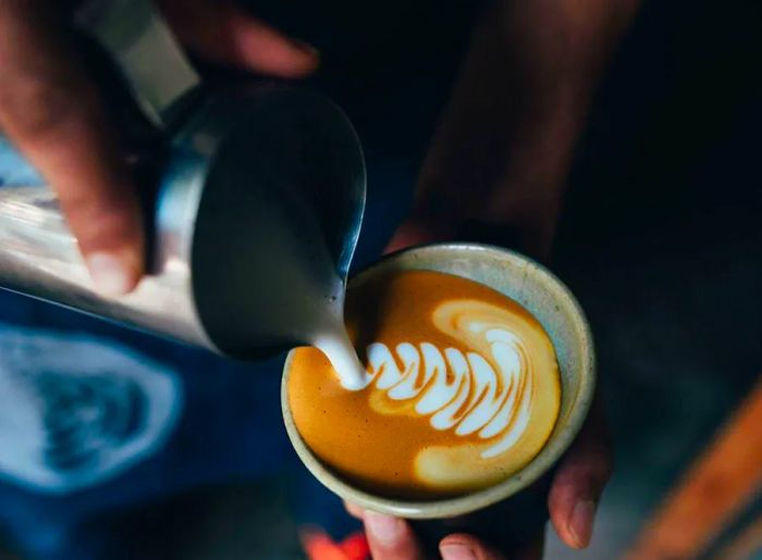 A close-up shot of a cappuccino in a ceramic cup as frothy milk is poured in by a barista.