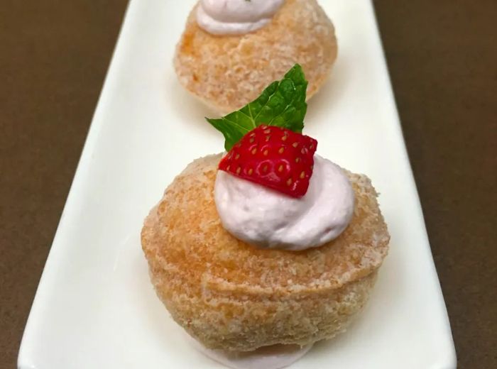 A tray filled with puffed pastries, each adorned with a dollop of whipped cream and a delicate slice of strawberry, gradually recedes into the background.