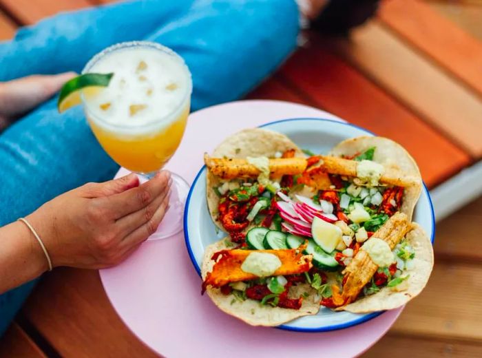 A vibrant pink side table holds a plate stacked with four tacos, while a woman relaxes nearby in a wooden lounge chair, sipping on a cocktail.