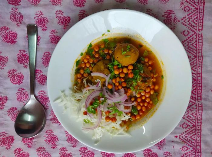 From above, a bowl of curry brimming with chickpeas, fluffy dumplings, fresh onions, herbs, and crisp cabbage, all resting on a beautifully patterned tablecloth.