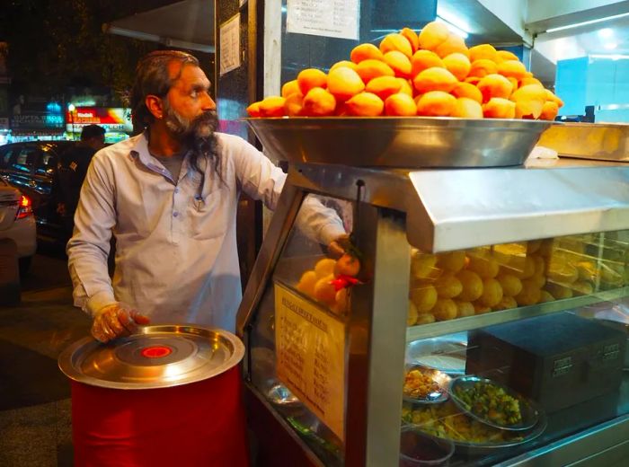 A vendor stands beside his cart, overflowing with trays of puffed, round golgappas.