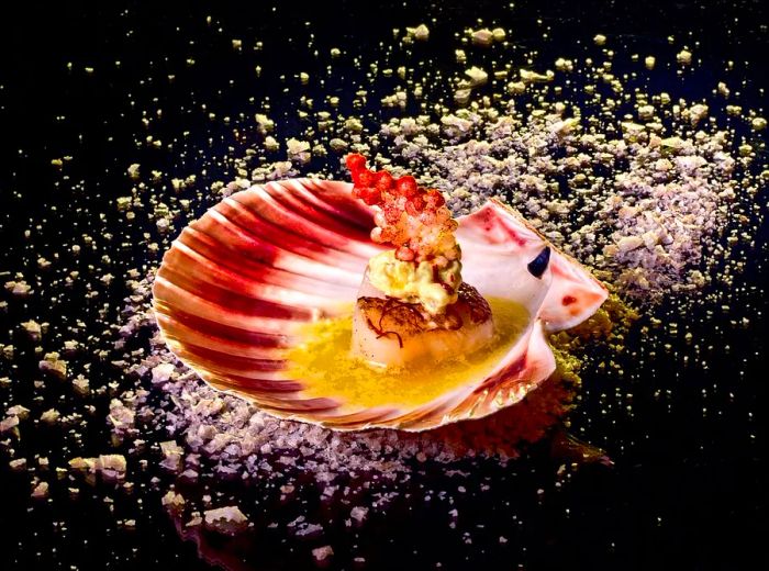 A scallop presented in its shell, topped with a slice of delicately balanced cauliflower, set against a minimalist backdrop.