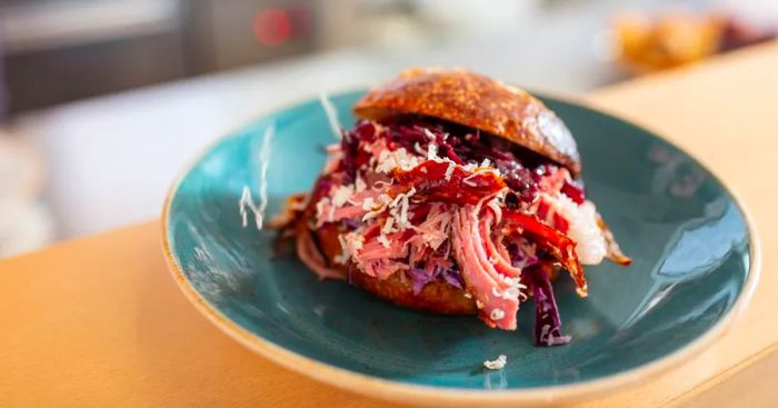 A hearty shredded meat sandwich bursting with meat, cheese, and cabbage, served on hamburger buns, rests on a ceramic plate atop a slender wooden counter