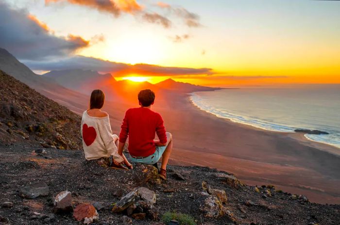 A young couple watches a stunning sunset from a mountain, overlooking the picturesque Cofete coastline on Fuerteventura island.