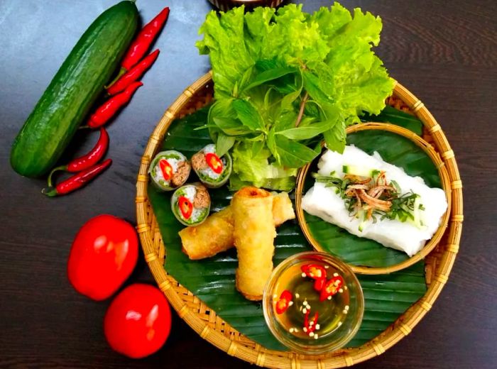 An aerial view shows a bamboo tray adorned with a leaf mat, showcasing an array of dishes including fresh greens, crispy rolls, and rice noodles topped with pork slices and aromatic herbs.