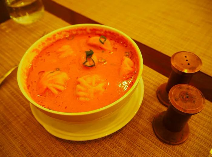 A bowl of dumplings floating in a rich tomato broth rests on a wooden table, with nearby condiment shakers ready for use.