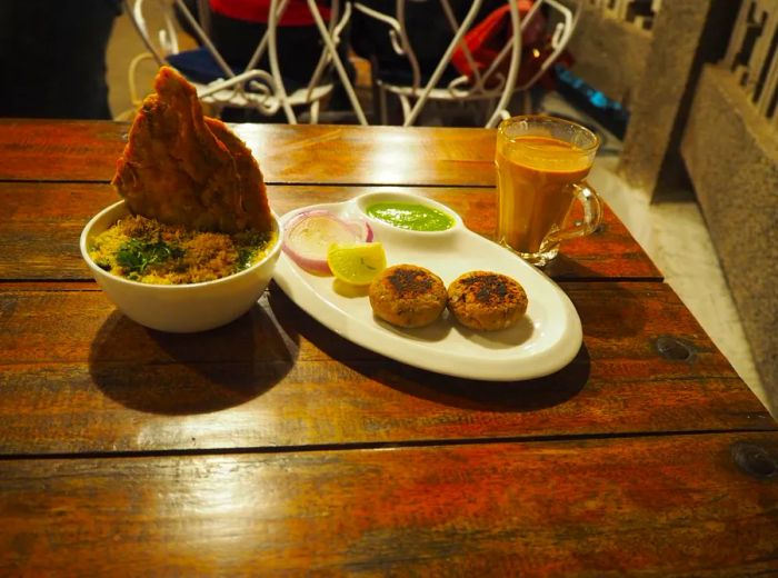 A wooden table outside the café is adorned with a spread of flattened kebabs with a dipping sauce, a bowl of crispy palak patta chaat, and a glass of steaming chai tea.