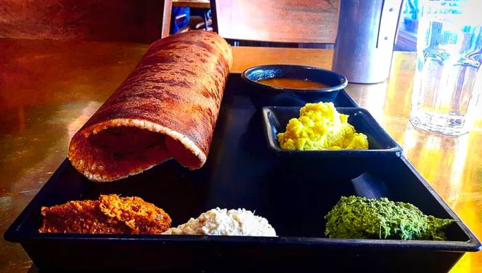 A tray showcasing a large rolled dosa accompanied by various small plates of chutneys and sides.