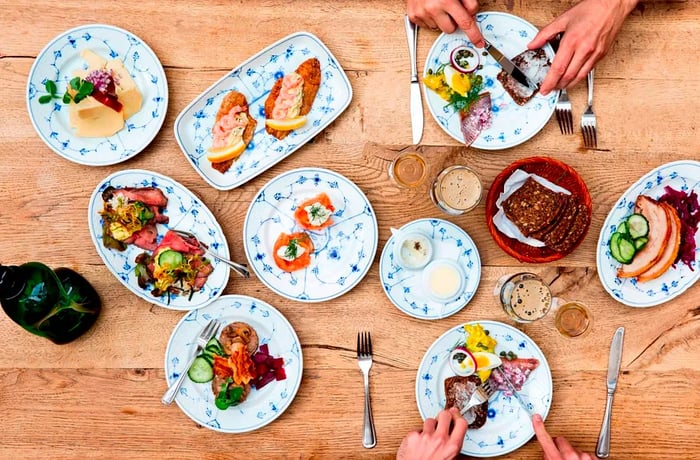 A top-down view of a wooden table adorned with an array of sandwich toppings including smoked fish, pickled vegetables, cold cuts, and various spreads, all arranged on decorative floral ceramic plates.