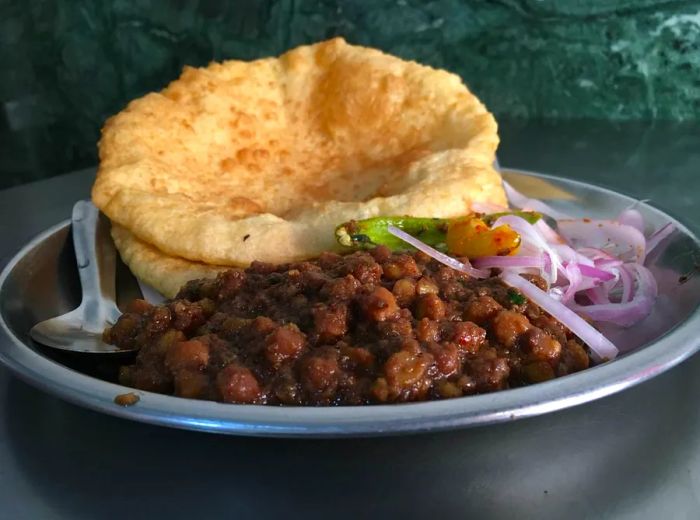 A vibrant display of chhole bhature features a mound of spicy chickpea curry paired with crisp, golden-brown flatbreads. The dish is surrounded by garnishes like sliced onions and fresh herbs, all set on a traditional platter with serving utensils.