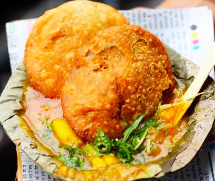 Two plump, deep-fried kachoris stand upright in a soupy paper bowl, with chunks of potatoes and sprigs of herbs floating in the rich stew.