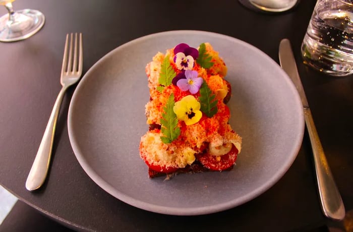 An open-faced sandwich lavishly adorned with layers of fish, grated cheese, and garnished with vibrant flowers and fresh leaves.