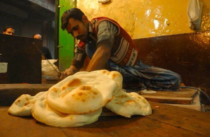 A stack of khameeri roti flatbreads is displayed on a wooden table, with a server working in the background against a light-colored interior.