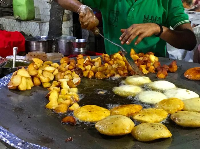 A sizzling griddle showcases chopped potatoes and airy kachoris, with a vendor busy mixing ingredients behind the hot plate.