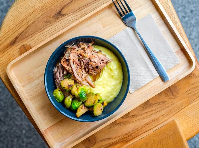 A top-down view of a wooden tray featuring mashed potatoes, shredded meat, and Brussels sprouts, accompanied by a fork and napkin