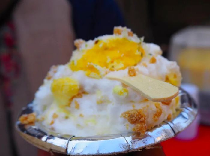 A serving of the creamy dessert known as daulat ki chaat, presented with a disposable wooden spoon on a paper plate.