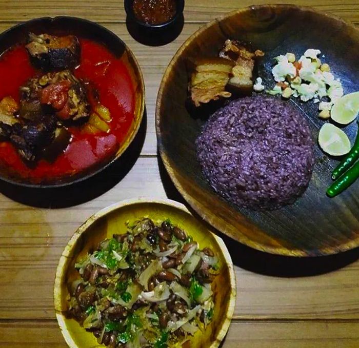 Viewed from above, a wooden table displays dishes including a serving of purple rice, slices of pork, and various garnishes.