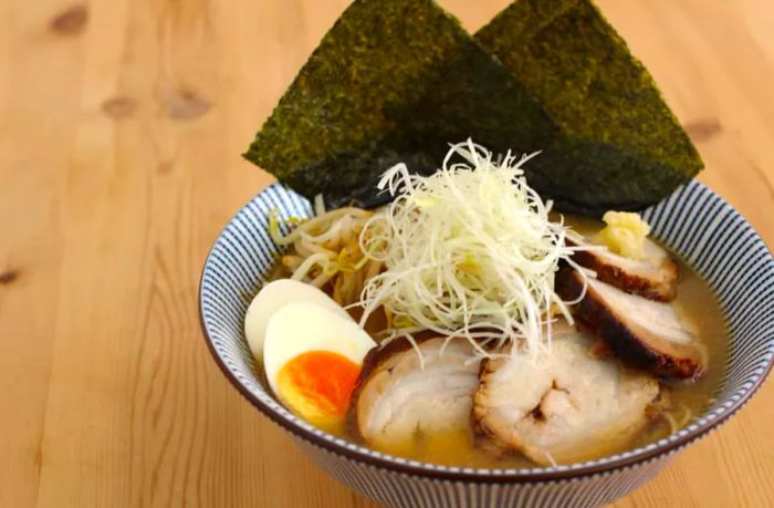 A beautifully arranged bowl of ramen on a wooden countertop, featuring pork slices, hard-boiled eggs, shredded cabbage, noodles, and nori sheets.