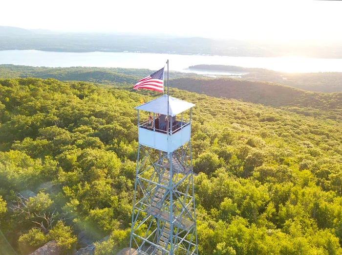 A woman stands in a tower, an American flag waving, gazing out over the Hudson River, lush greenery, and distant mountains.