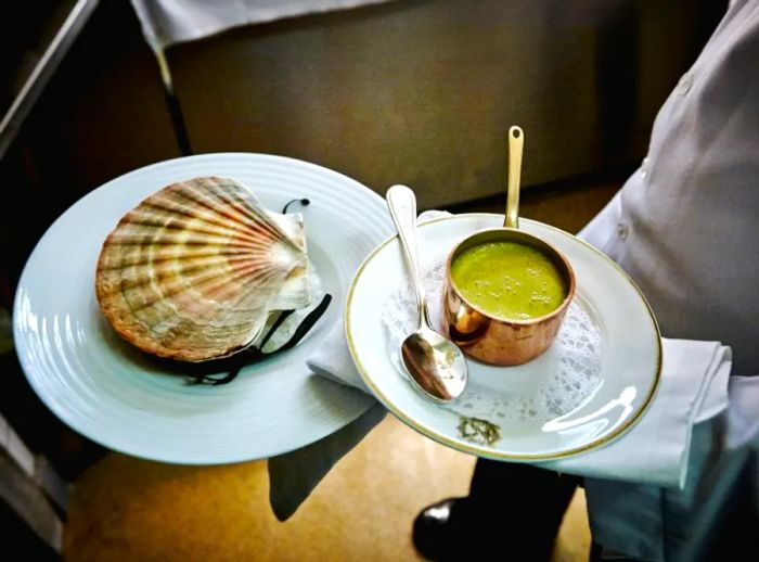 A server presents a plate featuring a large clam shell covering a dish, accompanied by a small copper saucepot and a serving spoon.