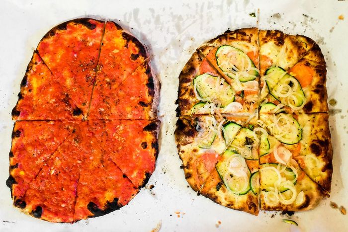 Aerial view of two New Haven-style thin-crust pizzas on white paper. One pizza has a classic tomato topping without cheese, while the other is a white pizza adorned with thinly sliced zucchini, onions, and basil, both featuring charred, slightly uneven crusts.