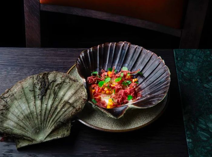 Sliced meat, creamy sauce, diced vegetables, and herbs presented in a clam shell against a dark wooden backdrop.