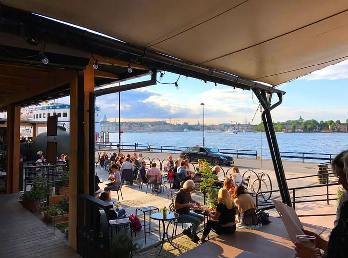 A bustling restaurant patio that stretches from sheltered awnings towards a boardwalk and the water beyond.