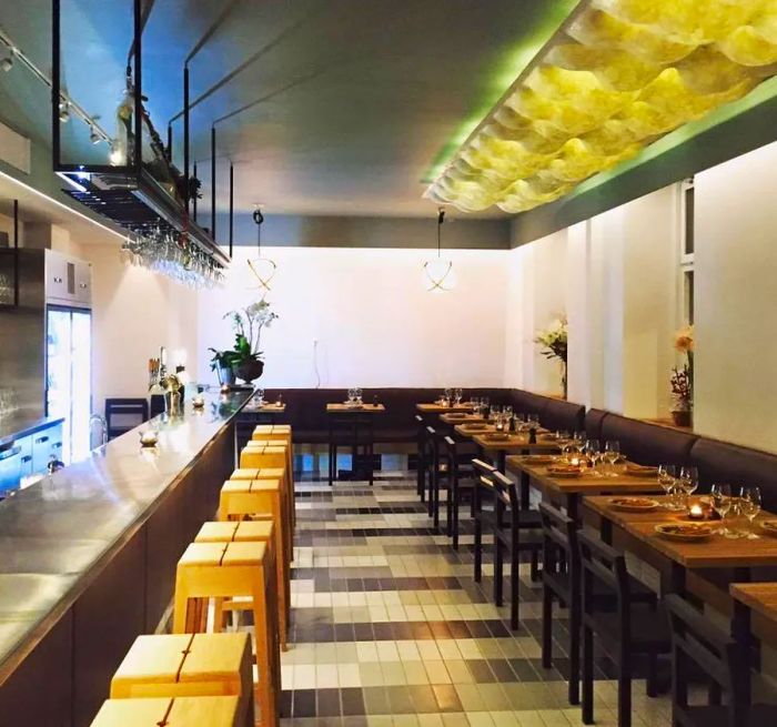 A bar counter lined with stools sits across from tables in a dining room featuring a stunning geometric ceiling installation.