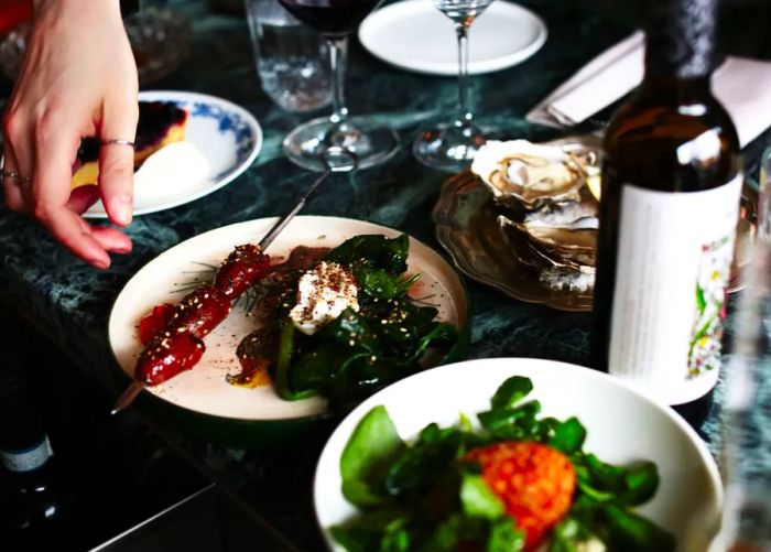 A server sets down a plate of meat skewers with yogurty greens on a dark marble table, accompanied by a salad, an oyster platter, and a bottle of wine.