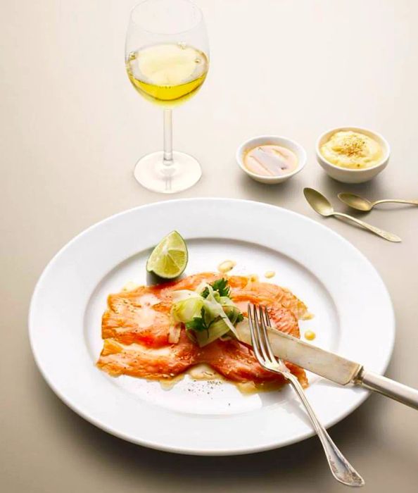 A fork and knife rest beside sliced salmon gravlax, accompanied by a lime wedge, a glass of wine, and various condiments.