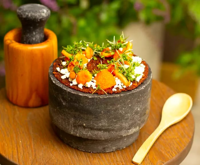 A rye pancake rests on a large stone surface, adorned with mushrooms, sour cream, shallots, and dill, accompanied by a mortar and pestle and a wooden spoon.