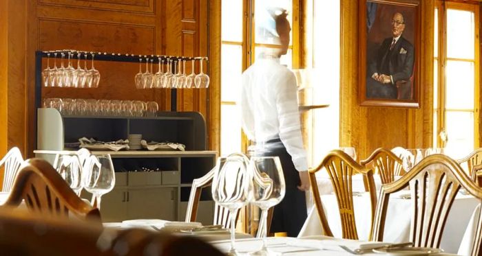 A server navigates through a dining room filled with elegantly set tables and bright, airy windows.