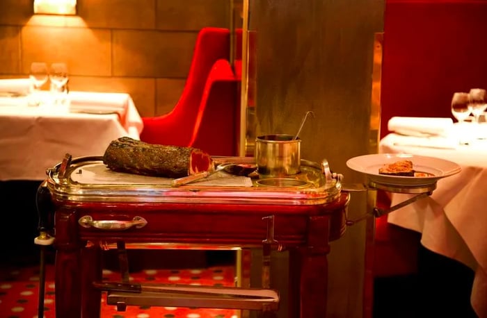 A dining room features a cart displaying a slab of cooked meat alongside serving utensils.