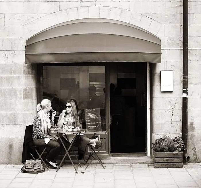 Two women enjoy a meal at a small patio table in front of a stone building with an awning.