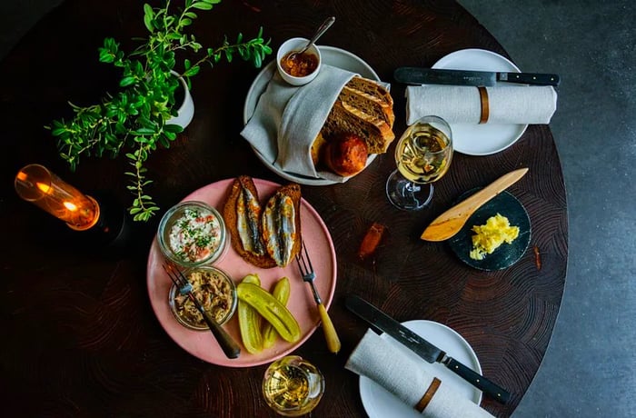 A bird's-eye view of a plate featuring sardines, pickles, and spread alongside a basket of bread and various other plates on a dark wooden table.