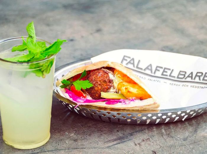 A pita sandwich filled with falafel and toppings, wrapped in branded wax paper, accompanied by a mint-garnished drink on a plate.