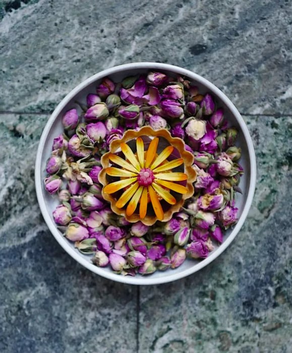 From above, yellow flower petals adorn a dish placed in a larger bowl filled with flower buds, all set against a marble backdrop.