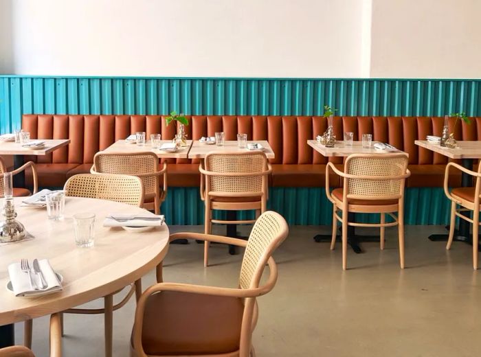 A cozy brown leather banquette positioned against teal walls in a bright, spacious dining area, featuring four and two-top tables, curved patio-style chairs, and neatly arranged table settings.