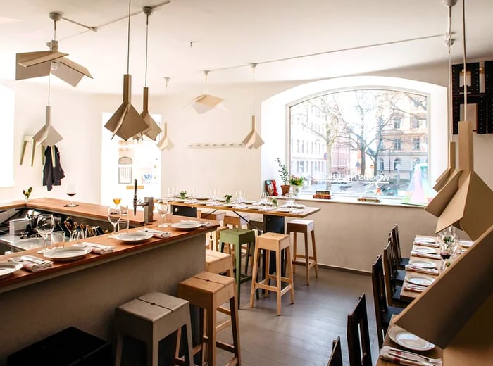 A bright dining area illuminated by sunlight, featuring a bar on one side and light wood tables lining the walls, adorned with geometric pendant lights.