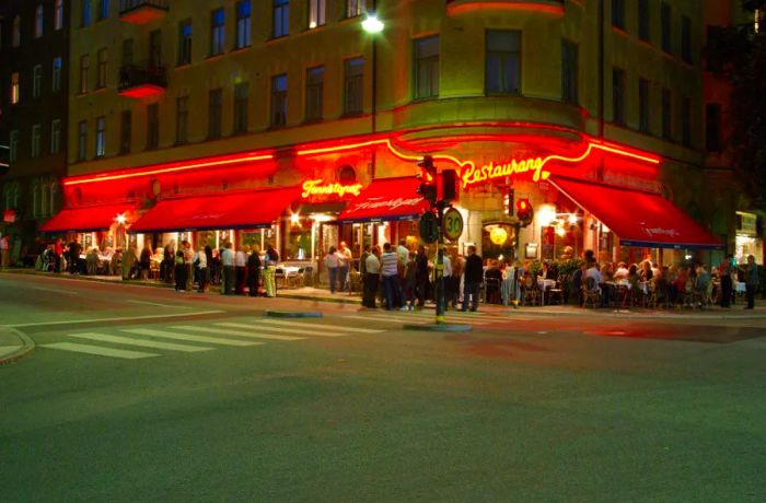 A bustling crowd gathers outside a corner eatery adorned with wide red awnings and bright neon signage reading Tennstopet.