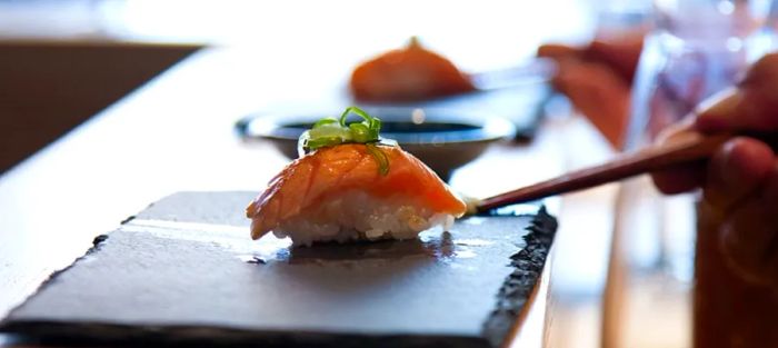 A beautifully crafted piece of sushi, adorned with a small dollop of greens, rests on a slate board with chopsticks nearby.