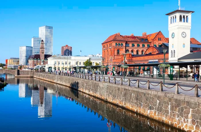 A riverside path with a stone wall leading to a canal, surrounded by a blend of historic and contemporary buildings.