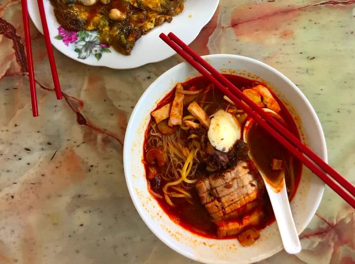 From above, a bowl of noodle soup enriched with pork in a savory broth, with a spoon and chopsticks perched inside, alongside a plate of omelet served with dipping sauce and more chopsticks, resting on a concrete surface.