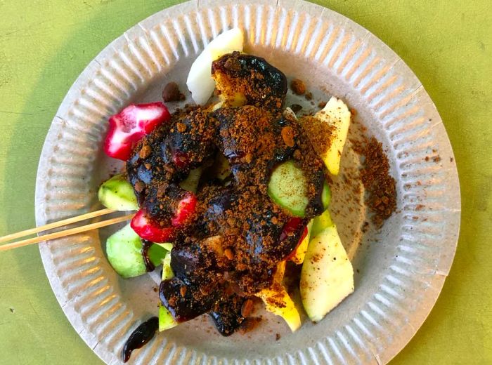 From above, a paper plate sits on a green plastic table, heaped with slices of fruits and vegetables covered in a thick, dark dressing, topped with a sprinkle of crushed nuts.