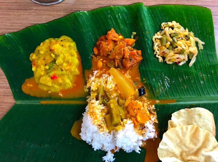 An overhead view of a partitioned banana leaf showcasing small portions of various dishes placed in designated sections, with a mound of rice at the center and two steel sauce canisters nearby.