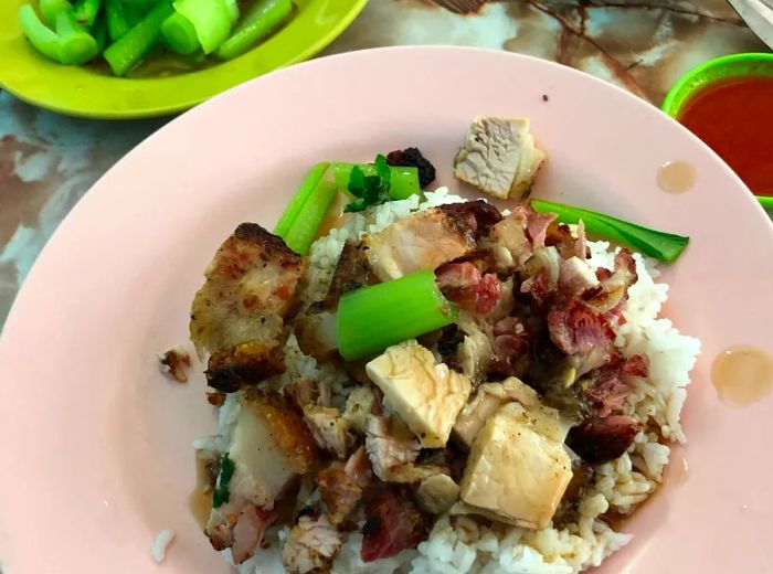 A slice of roasted pork resting atop rice, accompanied by greens on a pink plate, with a small plastic dish of additional greens and two cups of dipping sauce, all set against a colorful marble-like background.